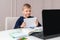 Boy shows notebook with answer to teacher on laptop screen. Smart Little Boy Is doing His Homework in Living Room. COVID-19