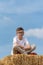 Boy sitting on hay bale with close eyes on blue sky background in summer. Carefree childhood. Vertical frame