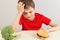 Boy at the table chooses between hamburger and broccoli on white background