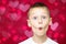 Boy teenager 7-10 in white t-shirt makes faces depicting kiss, with wide open eyes on red and pink hearts bokeh