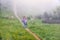 A boy traveler with hiking poles walks along the path on the green slope in heavy fog