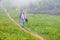 A boy traveler with hiking poles walks along the path on the green slope in heavy fog