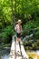 A boy traveler with a trekking poles stands on a rickety bridge over a fast stream