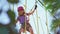 a brave kid girl in a helmet, purple T-shirt and shorts climbs in a rope park