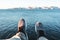 Brave traveler sitting on cliff and looks on legs and shoes. POV view on feet on background of ocean and mountains