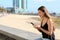 Brazilian young woman typing on phone sitting on wall with blurred city beach on the background. Copy space