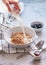 Breakfast, hand pours milk from a jug into muesli in a deep dish. Next to blueberries and jam on a blue-gray concrete background