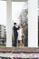 Bridal pair drinking champagne from wine glasses, bride and groom are between white columns outdoor