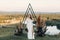 The bride with a bouquet of flowers stands in a triangular arch. Wedding ceremony in boho style.