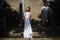 Bride in an elegant wedding dress stands on the background of a large staircase, and behind her is the groom in white clothes