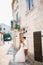 Bride and groom hug near a beautiful white building with balconies and wooden shutters in the old town of Perast near a