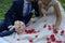 The bride and groom are sitting on a green meadow, strewn with red rose petals.