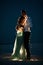 Bride and groom stand and hug in desert next to garland of light bulbs at night.