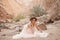 Bride in long dress and hat sits in canyon in sand.