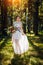 Bride in a long white dress stands in a clearing in the park among the trees with a bouquet of flowers in her hands. The bride