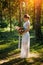 Bride in a long white dress stands in a clearing in the park among the trees with a bouquet of flowers in her hands. Summer
