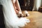Bride putting on white shoes on her nice feet. Sitting woman in white wedding gown. Wedding shoes on wooden floor.