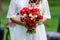 Bride in white dress holding in hands delicate, expensive, trendy bridal wedding bouquet of flowers in marsala and red colors. Clo