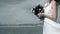 Bride in white dress is standing on ocean with bunch of flowers in her hands.