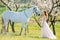 Bride in white dress stands near horse in spring blooming garden