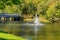 A bridge across the river with trees growing on the shore with yellow foliage and a fountain with clear streams of water