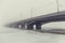 Bridge in fog over frozen river in ice and snow. Mysterious urban landscape in foggy day, vintage toned