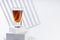 Bright amber brandy cocktail in shot glass in sunlight on white square podium as stairs with diagonal abstract stripes in soft.