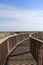 Bright blue skies over long wooden pier that stretches out over sandy dunes, leading one to the beach beyond