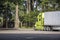 Bright green big rig industrial semi truck with refrigerator semi trailer running on the rest area parking lot with old trees to