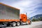 Bright orange classic American idol big rig semi truck with chrome parts transporting cargo in same color reefer semi trailer