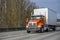 Bright orange day cab big rig semi truck with box trailer running on the wide multiline highway road with trees on the side