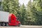 Bright red big rig long haul semi truck with refrigerator semi trailer moving on the road with green trees on the side