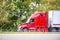 Bright red big rig semi truck with dry van semi trailer running quickly on the highway with green trees on the side for timely