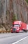 Bright red big rig semi truck with semi trailer running on winding mountain road with high cliff on the side