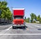 Bright red local moving and delivery rig semi truck with long box trailer standing on the crossroad on the city street