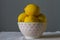 Bright ripe yellow lemons in a white porcelain bowl with pink balls in drops of water on a baggy cloth on a gray background.