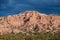 Bright sunlight illuminates a colorful red rock desert peak and badlands under dark, dramatic storm clouds near Santa Fe, New