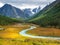 Bright sunny autumn landscape with winding river and  sunlit gold valley with green fir trees on mountainside under cloudy sky.