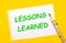 On a bright yellow background, a large wooden pencil and a white sheet of paper with the text LESSONS LEARNED
