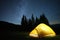 Brightly illuminated camping tent glowing on campsite in dark mountains under night stars covered sky. Active lifestyle