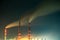 Brightly illuminated coal power plant high pipes with black smoke moving upwards polluting atmosphere at night with reflections of