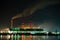 Brightly illuminated coal power plant high pipes with black smoke moving upwards polluting atmosphere at night with reflections of