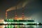 Brightly illuminated coal power plant high pipes with black smoke moving upwards polluting atmosphere at night with reflections of