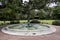 A bronze statue of a woman flying a flute in the center of a pond with weeping willow trees, lush green grass and plants