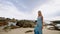 Bronzed woman strolls along sandy shore near sea. Elegance in blue gown against rocky backdrop, serene beach walk