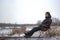 Brooding man sit on camping chair by frosty river in november, cold cloudy overcast weather with first snow