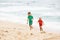 Brother and sister running, playing with sand and water on a tropical beach, dressed in protective wetsuit