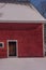 Brown barn door against the bright red clapboards against a snowy white field on a bright winters day.