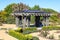 A brown and black pergola in the garden surrounded by a dirt path and lush green trees and plants