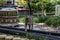 A brown cat walking over miniature train tracks surrounded by tiny homes and lush green trees, grass and plants